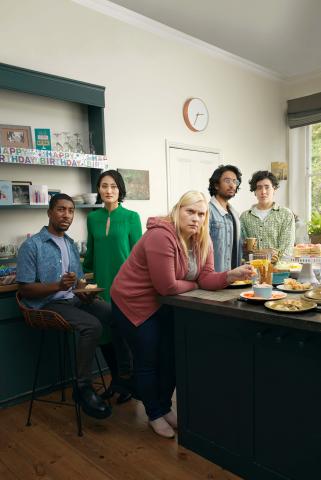 Two men and three woman are in a kitchen diner. They have expressions of concern and disbelief on their faces in reaction to something they've witnessed.
