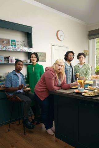 Two men and three woman are in a kitchen diner. They have expressions of concern and disbelief on their faces in reaction to something they've witnessed.