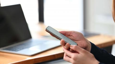 Hand holding mobile phone, laptop on desk in background. 